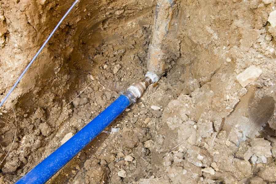 A close-up of a blue PEX pipe connected to an underground metal fitting as part of a residential main water line installation.